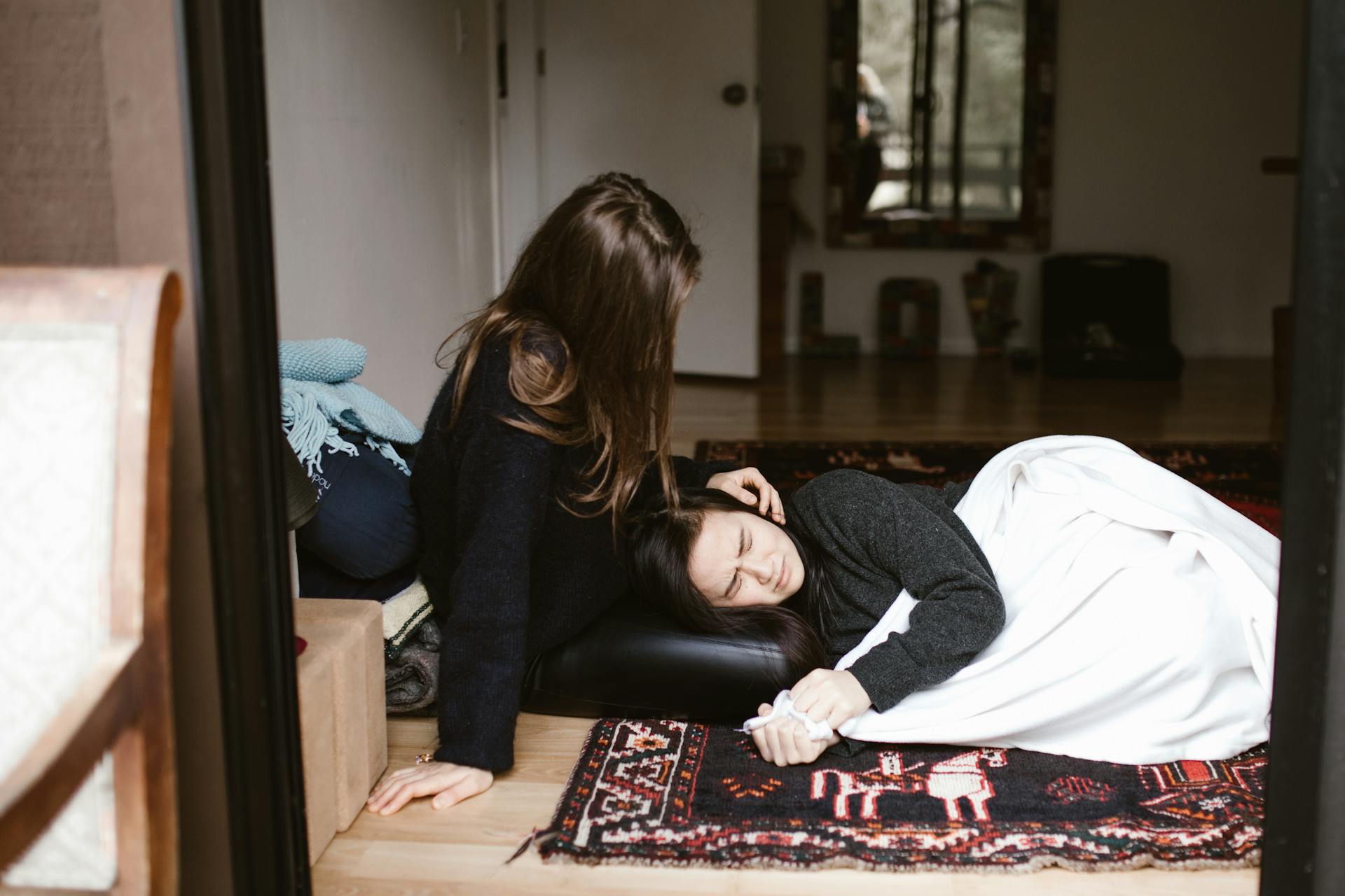 A woman sitting on the floor, comforting another woman who is lying down and holding her hand.