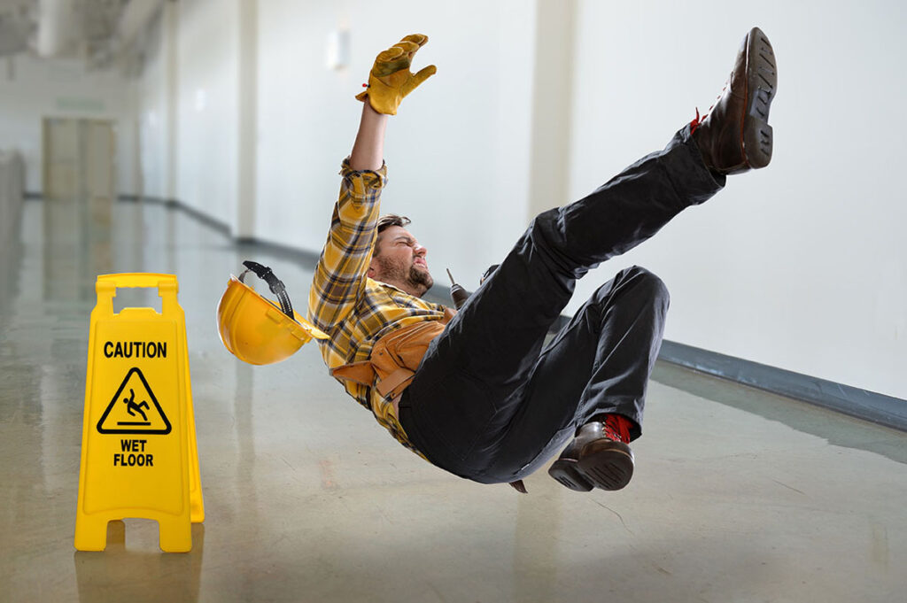 A construction worker falling backward on a slippery floor near a "Caution: Wet Floor" sign.