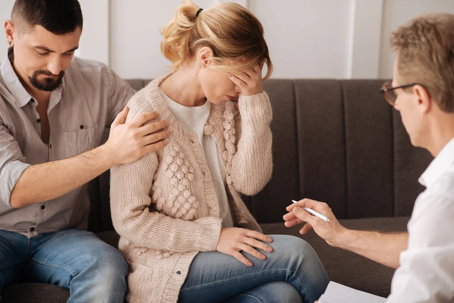 A man consoling a distressed woman while a therapist speaks to them.